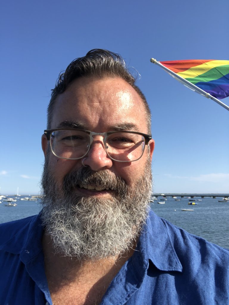 selfie of Kenneth on the deck of the Boatslip at tea dance with a pride flag and the Provincetown harbor in the background